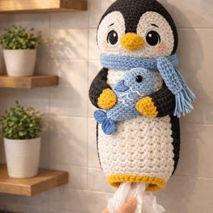 May include: A crochet penguin plastic bag dispenser featuring a black and white body, a yellow beak and feet, and a light blue scarf. The penguin holds a small blue fish in its wings, and a white plastic bag is visible at the bottom opening.