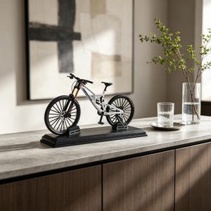 May include: A miniature white mountain bike model with black wheels and handlebars, mounted on a rectangular black display base. The frame features blue lettering and a rear suspension component, showcasing a detailed replica of a full-suspension bicycle.