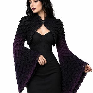May include: A black and purple ombre crochet shrug featuring a textured scale-like pattern, long flared bell sleeves, and a small decorative butterfly-shaped clasp at the neckline, worn over a simple black sleeveless dress.