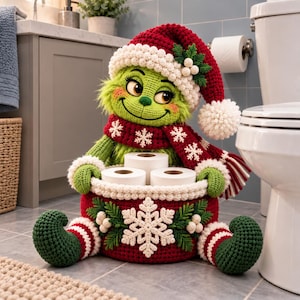 May include: A crocheted green creature doll wearing a red and white Santa hat and scarf, sitting and holding a round red basket decorated with a large white snowflake, green leaves, and white berries, containing three rolls of toilet paper.