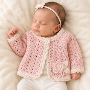 May include: A pale pink crocheted cardigan featuring a scalloped cream-coloured trim, a single floral button closure at the neckline, and a large decorative crocheted flower appliqué on the lower front. The garment has long sleeves and a textured pattern.