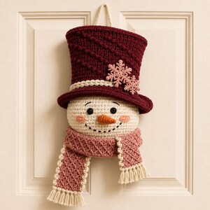 Könnte beinhalten: Ein gehäkelter Schneemann als Wandbehang mit einem dunkelroten Zylinder, der diagonale Texturstreifen und zwei hellrosa Schneeflocken-Applikationen aufweist. Das Gesicht ist cremefarben mit schwarzen Knopfaugen und einer orangefarbenen Nase.