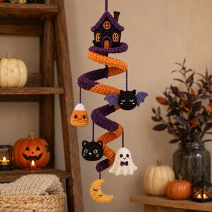 Könnte beinhalten: Ein hängendes Häkel-Mobile mit einer violetten und orangefarbenen Spiralbasis. Daran befestigt sind kleine gehäkelte Figuren, darunter ein schwarzes Spukhaus, eine Fledermaus, ein Zuckermais, ein schwarzes Katzengesicht, ein weißes Gespenst mit Fliege und ein gelber Halbmond.