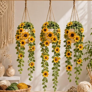 Könnte beinhalten: Drei hängende Häkel-Pflanzgefäße mit gelben Sonnenblumen mit braunen Mitten und grünen Blättern. Jeder Topf ist mit brauner Schnur an einer Holzstange befestigt. Die Häkelarbeit umfasst herabhängende Ranken mit kleinen Sonnenblumen und Blättern.