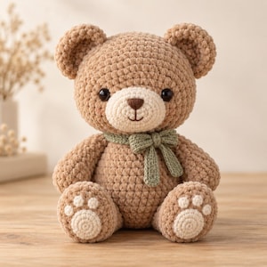 Könnte beinhalten: Ein hellbrauner, gehäkelter Teddybär mit cremefarbener Schnauze, dunkelbrauner Nase und schwarzen Augen. Der Bär trägt eine salbeigrüne, gehäkelte Schleife um den Hals und hat cremefarbene, kreisförmige Applikationen an den Fußsohlen.