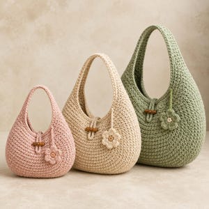 May include: Three crescent-shaped crochet handbags in graduated sizes, colored dusty pink, beige, and sage green. Each bag features a single handle, a central wooden toggle closure, and a small matching crocheted flower charm attached to the side.