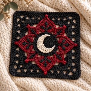May include: A square crochet motif featuring a central cream-colored crescent moon surrounded by a dark red floral pattern with black accents. The entire design is framed by a black crocheted border with an openwork lattice pattern throughout.
