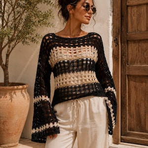 Könnte beinhalten: Ein langärmeliger Häkelpullover mit einem schwarz-beigen Gittermuster, weiten Glockenärmeln und einem Rundhalsausschnitt, kombiniert mit einer hellbeigen Hose mit Kordelzug. Das Kleidungsstück hat eine durchgehende, offen gestrickte Textur.