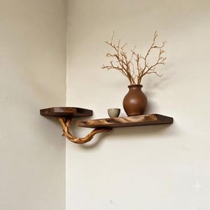 Op de afbeelding: Een hoekwandplank met twee niveaus van donker hout met natuurlijke randen. De planken worden ondersteund door een gebogen, lichtgekleurde tak. Een bruine keramische vaas met dunne, kale takjes en een kleine beige kom staan op de onderste plank.