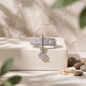 May include: A silver-toned ring featuring a wide band encrusted with small clear stones and a row of larger square-cut clear stones. A vertical bar extends from the center, holding a single round, dangling clear stone charm at the bottom.