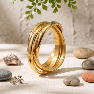May include: A gold-toned ring composed of three interlocking bands with a polished, reflective finish. The circular bands are connected in a continuous loop, creating a layered, dimensional appearance with smooth, rounded edges and a bright metallic sheen.