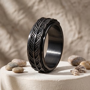 Puede incluir: Un anillo de metal negro mate que presenta un patrón de plumas en relieve continuo alrededor de la banda exterior. El anillo tiene un perfil ancho y plano con un acabado oscuro cepillado y una superficie interior lisa con detalles tallados.