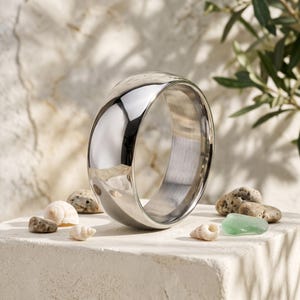 May include: A polished silver-toned metal ring with a smooth, domed exterior surface and a brushed finish on the interior band. The ring is positioned upright, showcasing its reflective metallic sheen and rounded profile in a clear, simple design.