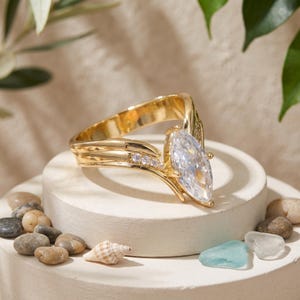 May include: A gold-toned ring featuring a large marquise-cut clear stone as the centrepiece, accented by a row of small round clear stones along the band. The band has a split, V-shaped design that tapers towards the back of the ring.