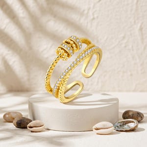 May include: A gold-toned open ring featuring two distinct bands. One band is a smooth, textured wire with a central rotating bead element, while the second band is encrusted with a row of small, clear, sparkling stones along its outer surface.