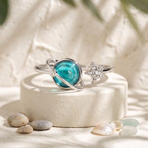 May include: A silver-toned open ring featuring a spherical teal blue stone encased in a thin metal ring resembling a planet on one end and a small, five-pointed star encrusted with clear, sparkling stones on the opposite end of the band.