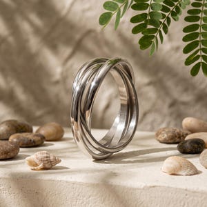 Può includere: Un anello color argento composto da tre fasce intrecciate che creano un anello fluido e continuo. La superficie metallica è liscia e riflettente, catturando la luce per evidenziare il profilo arrotondato della struttura circolare.