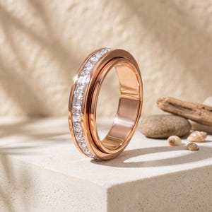 May include: A rose gold-toned ring featuring a central channel set with a continuous row of clear, rectangular-cut stones. The band has a polished, smooth finish and a rounded profile, showcasing a metallic luster and reflective, faceted gems.