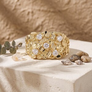 May include: A gold nugget ring featuring a textured, irregular surface with five round, clear, faceted stones embedded across the front. The band has a wide, chunky design with a polished finish that reflects light, creating a high-contrast appearance.