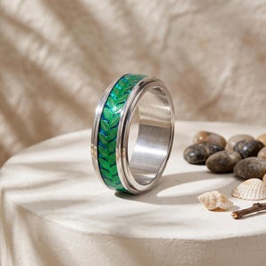May include: A silver-toned metal ring featuring a central band with a vibrant green leaf pattern set against a blue background. The ring has a smooth, polished finish on the edges and a wide, flat profile, showcasing a detailed botanical design.