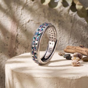 May include: A silver ring featuring a band with a repeating pattern of iridescent blue and purple leaf-like shapes. The interior of the band is engraved with text and the stamp 925, indicating sterling silver composition. The ring has an open design.
