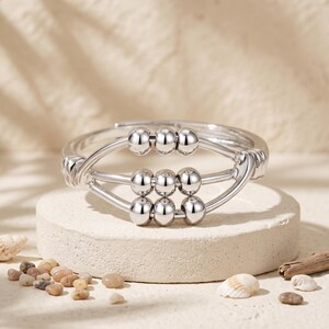 May include: A silver-toned ring featuring a central design of three horizontal bars, each holding three small, movable spherical beads. The band is open at the back, and the metal has a polished, reflective finish throughout the entire structure.