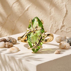 May include: A gold-toned ring featuring a large, marquise-cut, translucent lime-green gemstone. The setting includes a metallic dragonfly perched on the upper left side of the stone and a curved, leafy vine branch extending along the right side.