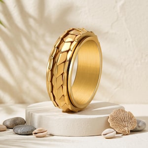 May include: A gold-toned ring featuring a textured outer band with a repeating scale-like pattern. The ring has a smooth, polished interior surface and a metallic finish, designed with a wide band profile that showcases intricate geometric detailing.