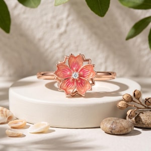 Könnte beinhalten: Ein Ring aus Roségold mit einem fünfblättrigen Blumendesign und Farbverlauf in Pink und Orange aus Emaille. In der Mitte der Blüte befindet sich ein kleiner, klarer, runder Edelstein. Die Ringschiene besteht aus glattem, poliertem Roségold.