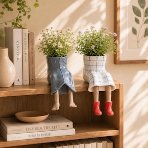 May include: Two ceramic planters shaped like seated figures with dangling legs. One planter is blue with a denim-like texture, and the other is white with a blue grid pattern. Both contain small green plants with white and purple flowers.