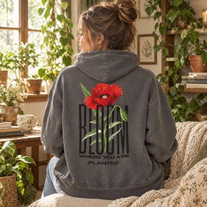 Peut inclure: Un sweat à capuche gris anthracite avec un grand motif de coquelicot rouge au dos. Le mot "BLOOM" est imprimé en grandes lettres noires derrière la fleur, avec les mots "WHERE YOU ARE PLANTED" imprimés en plus petit texte noir en dessous.