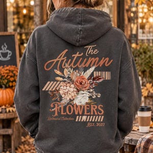 Peut inclure: Un sweat à capuche gris anthracite avec un motif au dos portant l'inscription The Autumn FLOWERS Botanical Collection EST. 2027. Le design présente un arrangement floral central composé de roses et de feuillage séché dans des tons orange et crème.