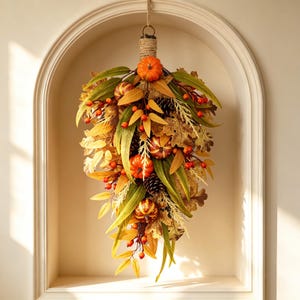 May include: A teardrop-shaped autumn swag featuring orange pumpkins, brown pinecones, and red berries nestled among green and yellow foliage. The arrangement is bound at the top with natural twine, creating a decorative hanging piece.