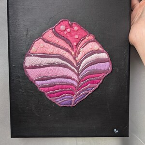 Puede incluir: Un lienzo negro cuadrado con una forma central bordada similar a una concha. El diseño utiliza puntadas de satén horizontales en tonos rosa, magenta y morado, acentuadas con pequeñas cuentas redondas de color rosa pálido y un borde brillante.