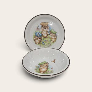 以下が含まれることがあります： ダークブラウンの縁取りがある白い陶器のボウル2つ。上のボウルには、帽子をかぶった3匹の茶色の犬が庭にいるイラストが描かれています。下のボウルには、緑色の帽子をかぶった茶色の犬が1匹、花や鳥のいる庭にいる様子が描かれています。