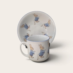 May include: A white ceramic mug and matching saucer set featuring a repeating illustration of a brown rabbit wearing a blue jacket. The mug has a curved handle and a thin blue rim, while the saucer displays multiple rabbits in various poses.