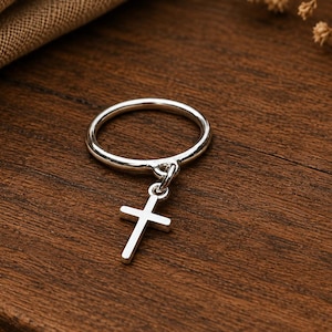May include: A polished silver-tone ring featuring a small, dangling cross charm attached to the band by a circular jump ring. The metal surface is smooth and reflective, showcasing a simple, clean design with a classic cross pendant element.