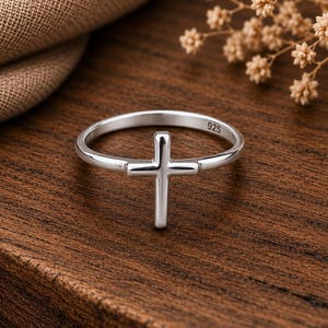 Peut inclure: Une bague en argent poli avec un motif de croix centré. L'anneau est fin et lisse, avec le nombre 925 clairement gravé sur la surface intérieure, indiquant une composition en argent sterling. La bague présente une finition métallique brillante.
