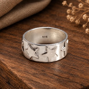May include: A silver band ring featuring a repeating embossed star pattern around the exterior. The interior of the ring is clearly stamped with the number 925, indicating the silver content. The metal has a polished finish with subtle texture.