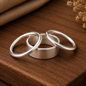 May include: A set of three silver rings consisting of two thin, smooth, polished bands and one wider, flat band. The interior of the wider band is clearly stamped with the hallmark 925, indicating the silver content of the jewellery pieces.