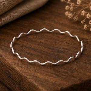 Può includere: Un bracciale rigido sottile di colore argento caratterizzato da un design continuo ondulato o a serpentina. Il metallo presenta una finitura liscia e riflettente che cattura la luce, creando una forma circolare semplice con un motivo ondulato costante.