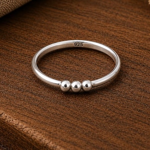 Peut inclure: Une bague en argent poli composée d'un anneau fin et lisse orné de trois petites perles mobiles en argent au centre. L'intérieur de l'anneau porte l'inscription 925, indiquant la composition en argent sterling de ce bijou.