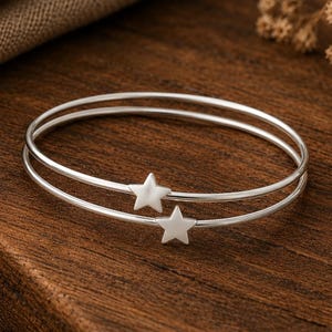 May include: A double-layered silver bangle bracelet featuring two small, polished star-shaped charms. The thin, circular bands are connected and crafted from a smooth, reflective metallic material, creating a simple and clean design.