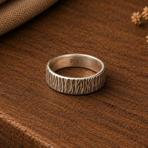 May include: A silver ring with a textured, bark-like exterior finish. The interior of the band is smooth and clearly stamped with the hallmark 925, indicating sterling silver composition. The ring features a consistent width and a metallic sheen.