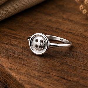 May include: A polished silver ring featuring a circular button-shaped centrepiece with four holes in the middle. The inner band of the ring is clearly stamped with the number 925, indicating the sterling silver composition of this jewellery piece.