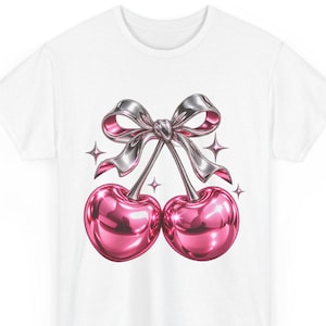 Può includere: Una t-shirt bianca con una grafica raffigurante due ciliegie rosa metallizzato collegate da un fiocco argento metallizzato. Il design include piccoli accenti a forma di stella a quattro punte attorno alle ciliegie.