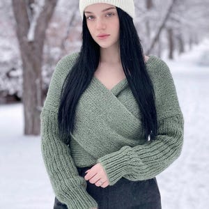 May include: A sage green long-sleeved knit jumper featuring a wrap-style front with a V-neckline and ribbed cuffs, paired with a solid white knit headband. The jumper has a chunky texture and a relaxed fit, while the headband is wide and ribbed.