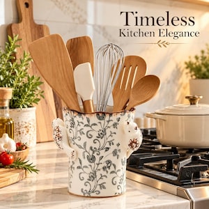 May include: A ceramic utensil holder with a white base and gray floral pattern, featuring two small bird figures on the sides. It contains wooden spatulas, a white silicone spatula, a metal whisk, and a wooden spoon. Text reads Timeless Kitchen Elegance.