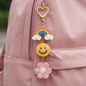 May include: A gold-toned heart-shaped clasp keychain featuring three crocheted charms: a multi-coloured rainbow with white clouds, a yellow smiley face with pink cheeks, and a light pink flower with a white centre, all linked by small gold chains.