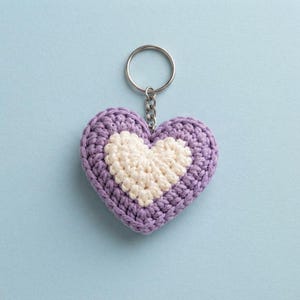 Peut inclure: Un porte-clés en forme de cœur avec un design en crochet bicolore. Le centre est un petit cœur de couleur crème entouré d'une bordure plus épaisse en fil violet clair. Un anneau fendu en métal argenté est fixé au sommet du cœur.